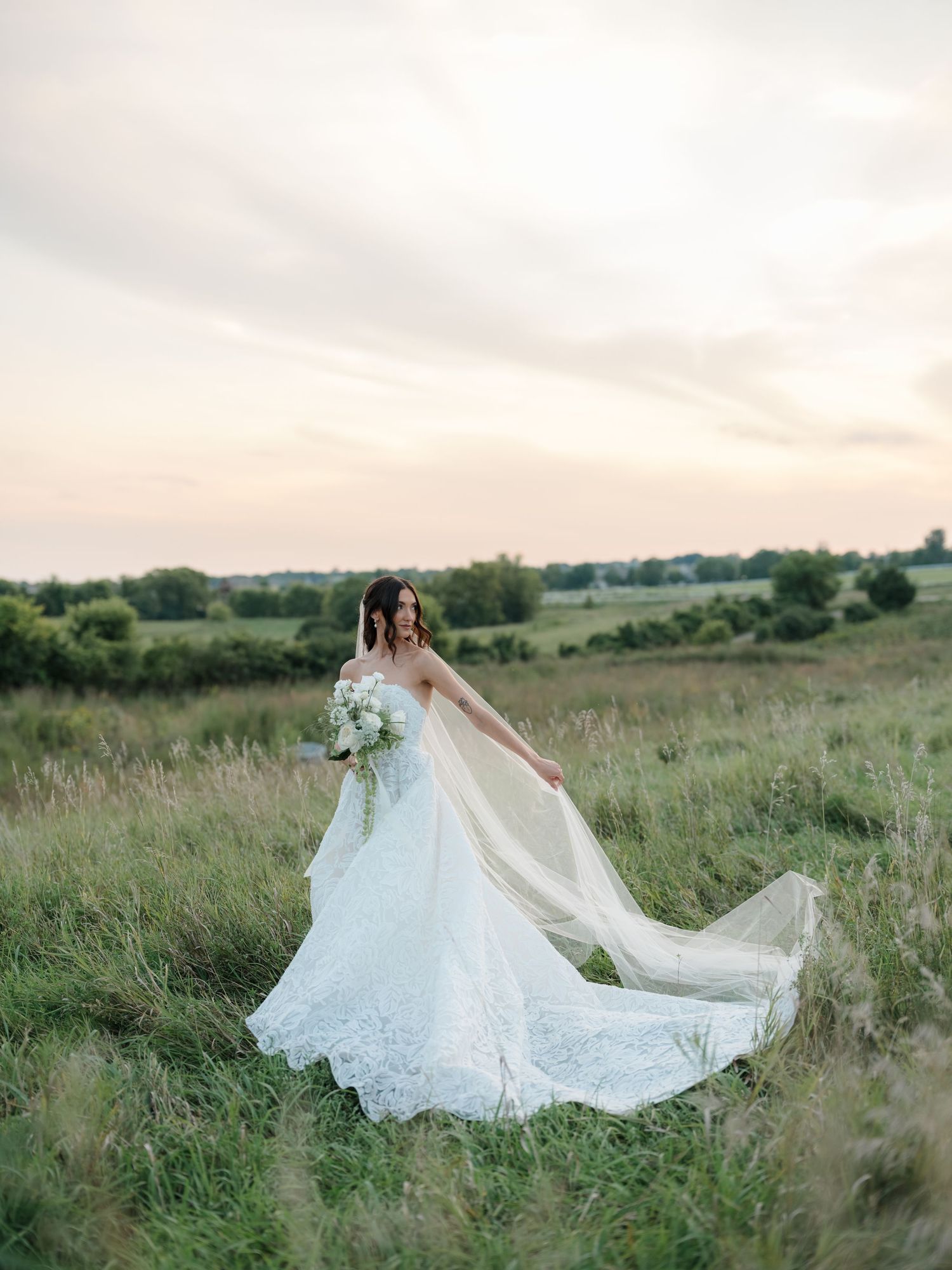 A bride in a flowing white lace gown poses gracefully in a sunlit meadow during golden hour.