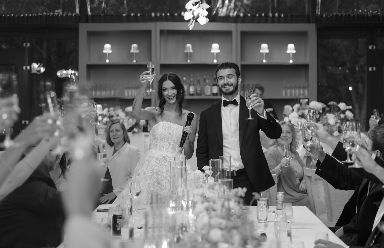 A joyful black and white photo of newlyweds standing and raising glasses during a wedding reception toast.