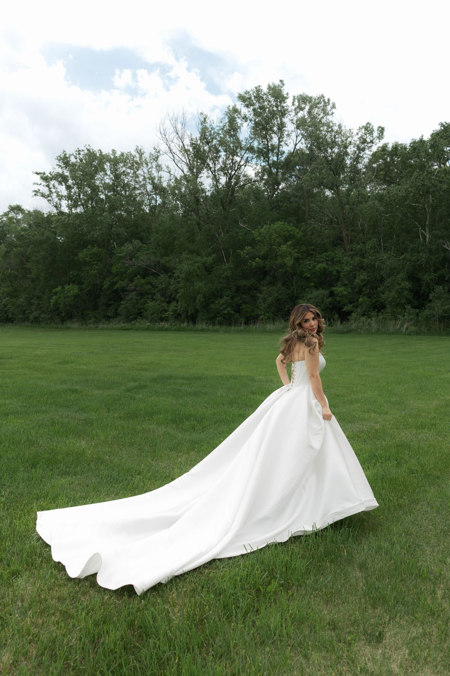 A flowing white wedding dress train sweeps dramatically across a lush green meadow with trees in the background.