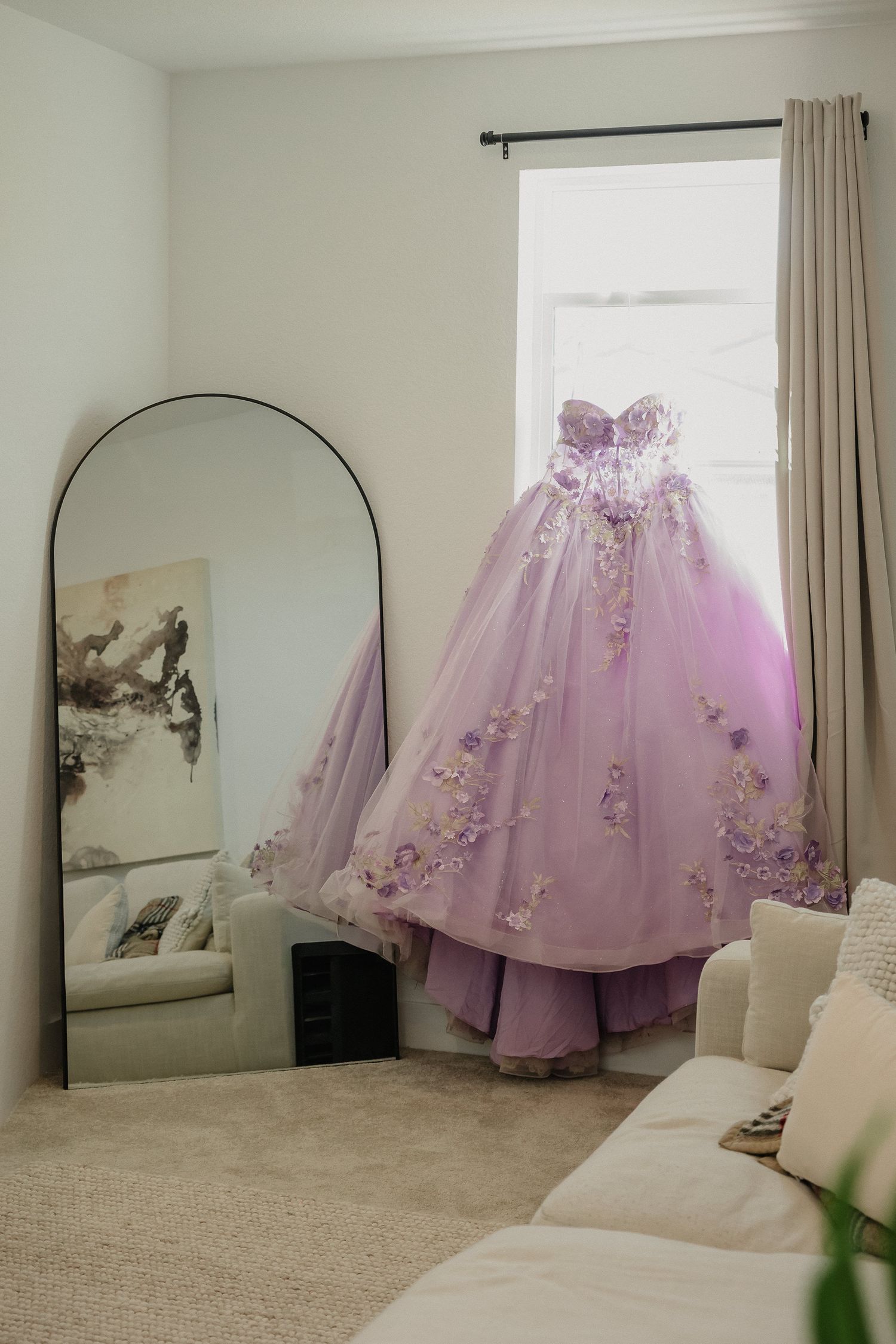 Lavender ball gown with floral details hangs elegantly in front of a full-length mirror in a bright room.