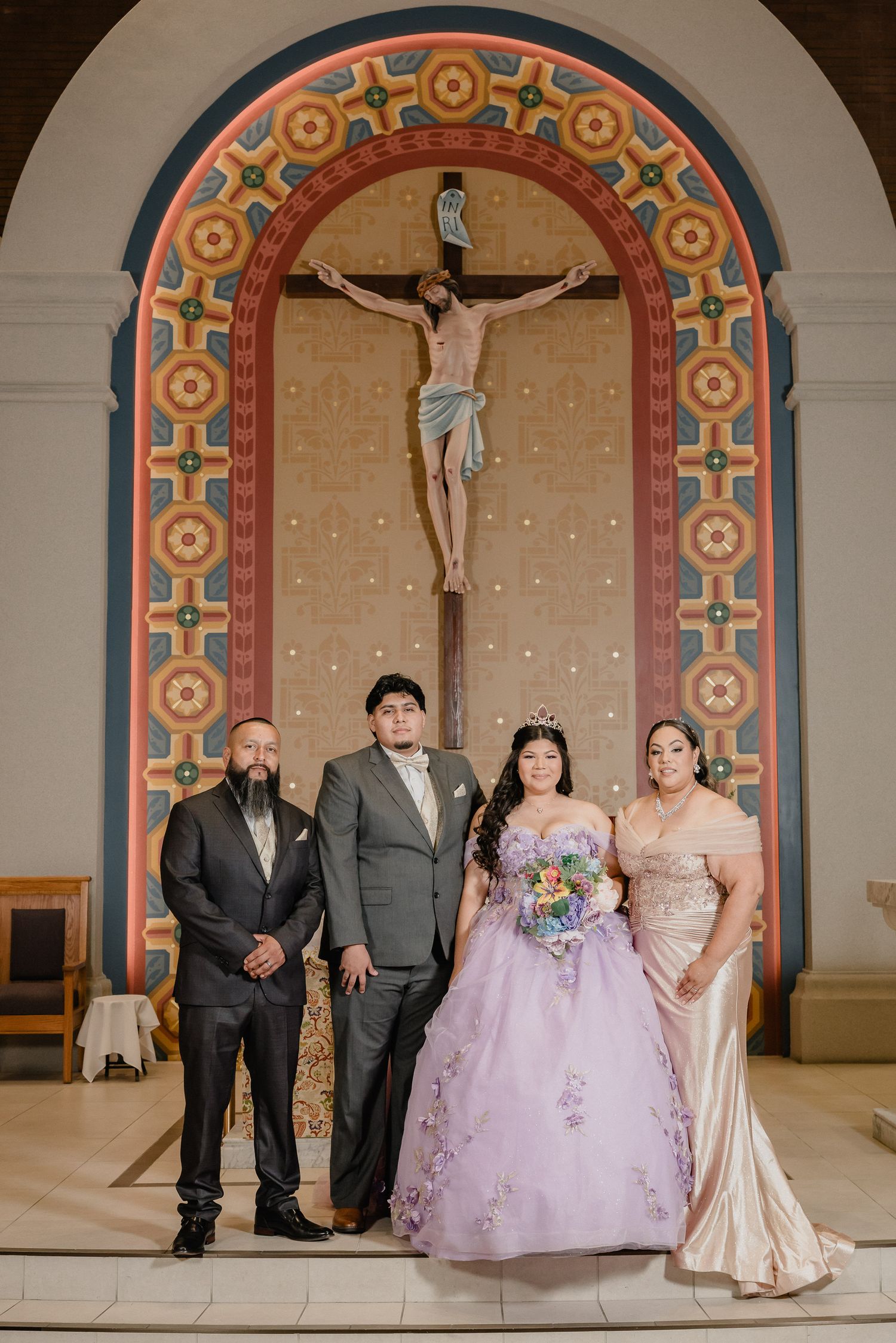 Wedding party poses in front of ornate church altar with decorative arches and crucifix.