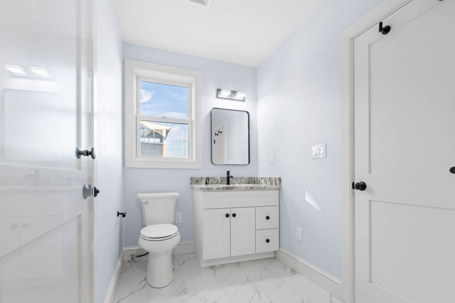 Clean white bathroom with marble tile floors, vanity with granite top, toilet, and natural light.