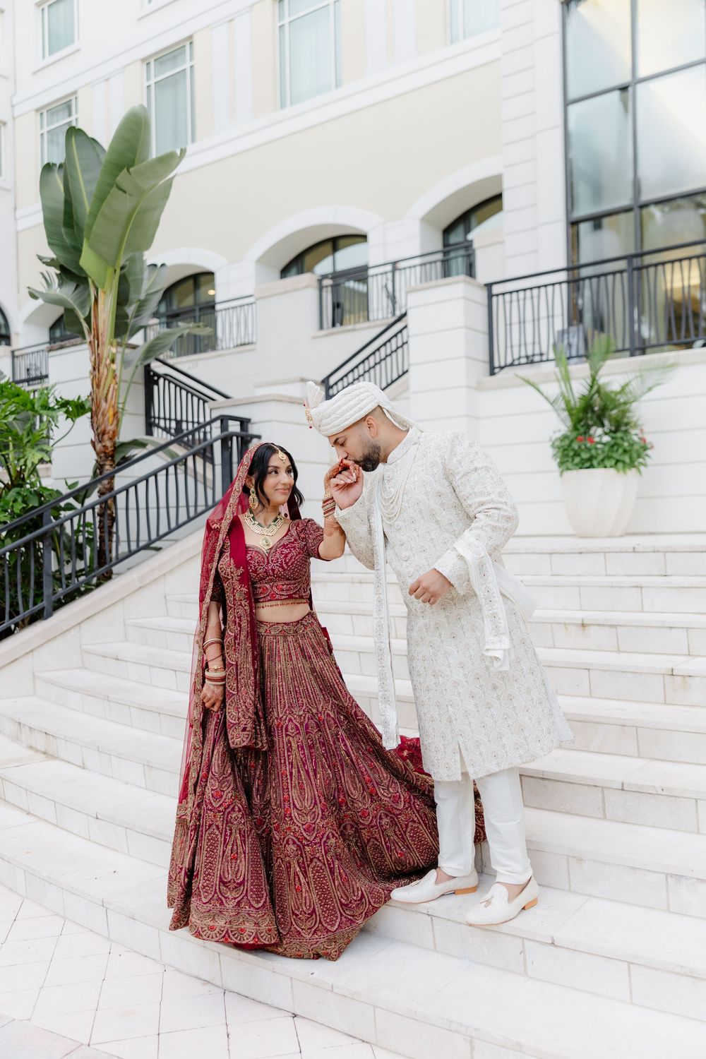 A couple in traditional South Asian wedding attire poses on elegant white marble stairs with tropical plants in the background.