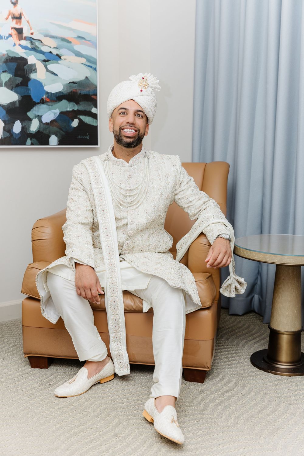 Person in all-white traditional Indian attire sits on tan leather couch with artwork visible in background.