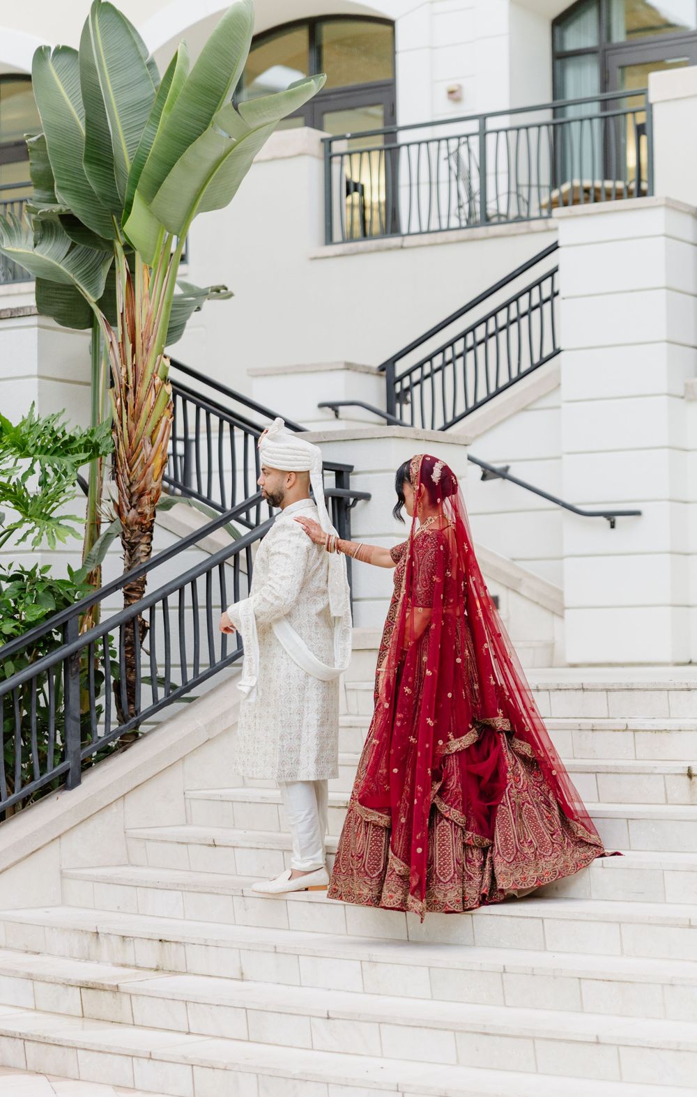 A couple in traditional Indian wedding attire walks up white marble stairs with ornate railings and tropical foliage.