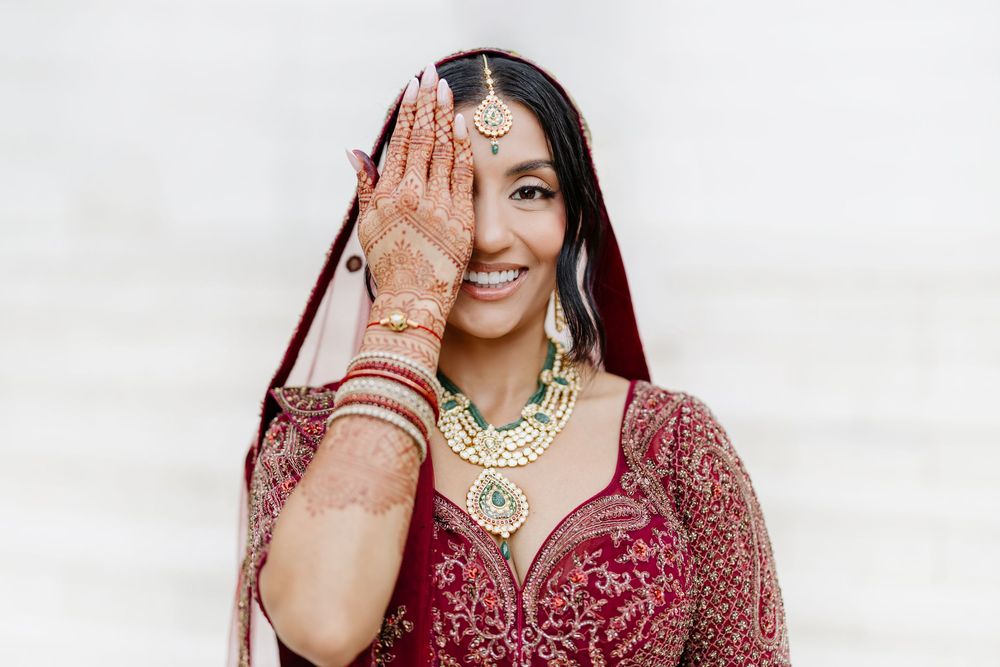 Traditional Indian bride in red lehenga with henna decorated hands and gold jewelry poses against white background.