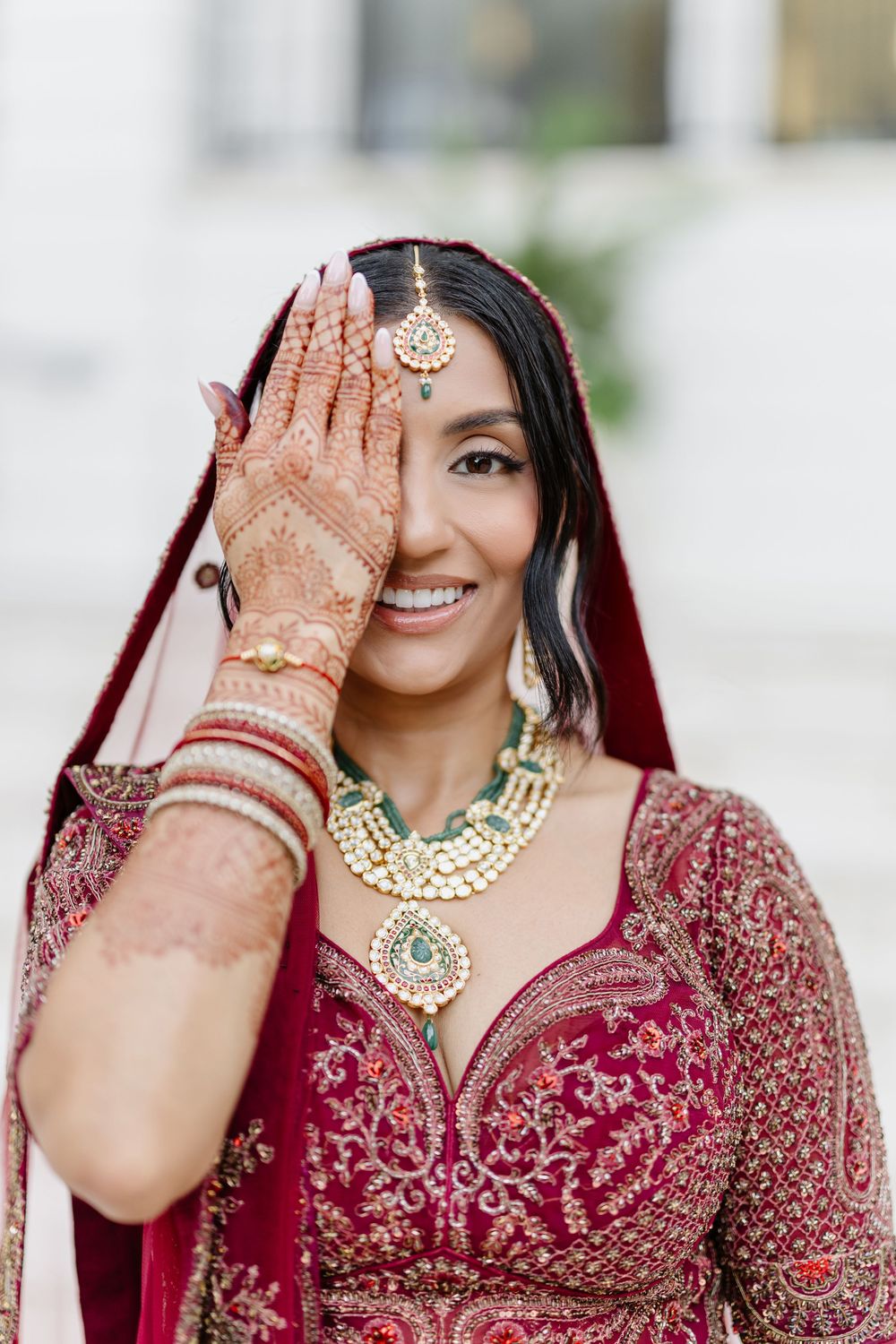 A bride in a burgundy lehenga with intricate embroidery shows off traditional henna designs and gold jewelry.