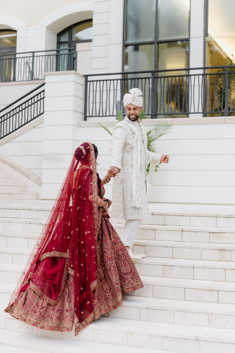 A couple in traditional South Asian wedding attire descends white marble stairs in an elegant outdoor setting.