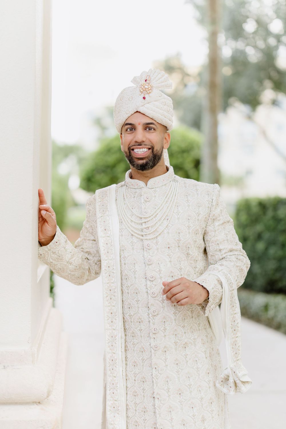 A groom in a traditional cream-colored sherwani and turban poses elegantly against a white pillar outdoors.