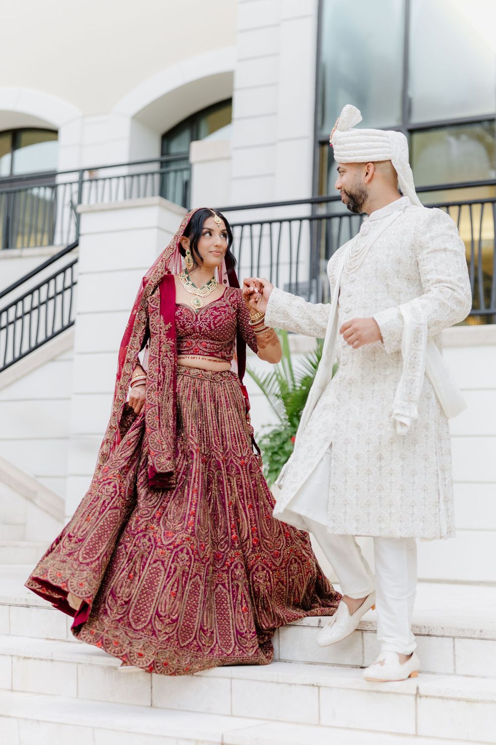 A couple in traditional South Asian wedding attire sits together on white marble steps during their wedding photoshoot.