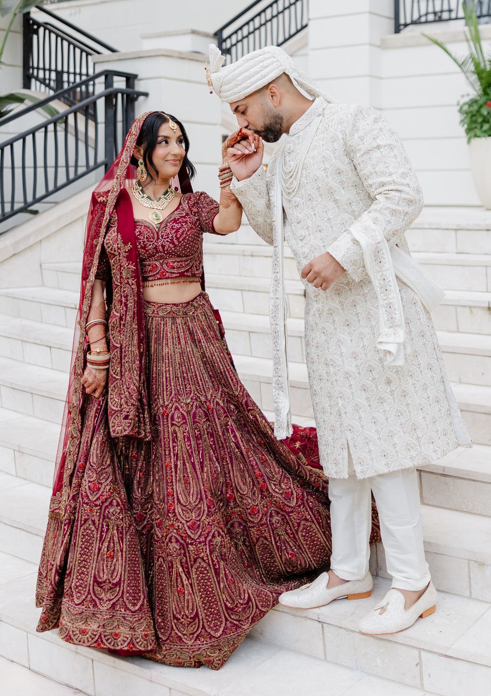 A couple in traditional South Asian wedding attire poses on white marble stairs, with burgundy and gold lehenga details.