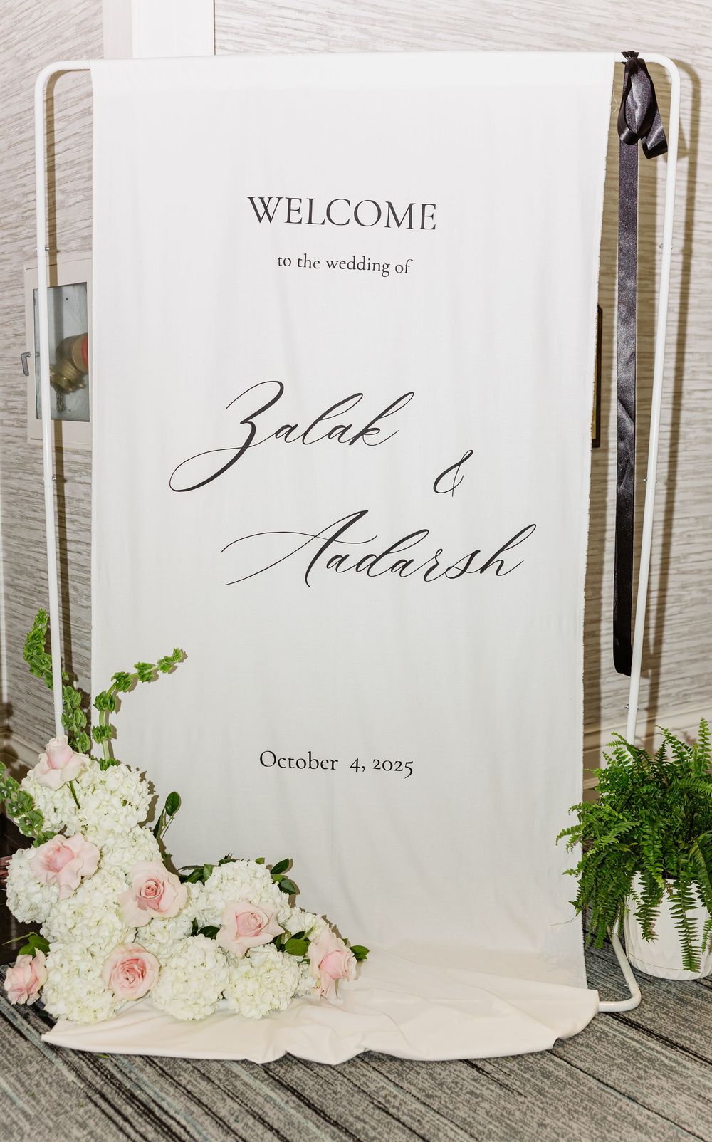 Elegant wedding welcome sign with calligraphy text and soft pink and white floral decorations against white backdrop.