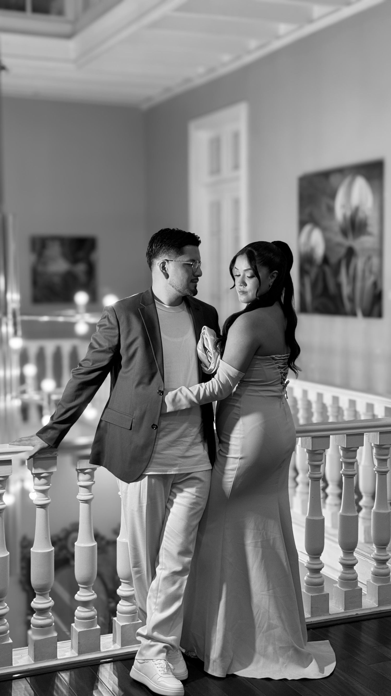 Romantic couple in formal attire standing by white balustrade in black and white photography.