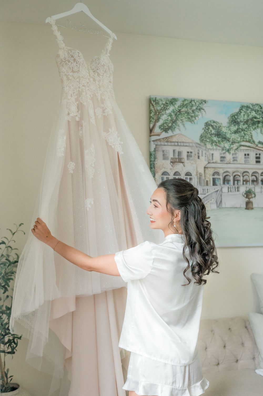 A white lace wedding dress hangs in a room with a decorative wall painting as someone admires it.