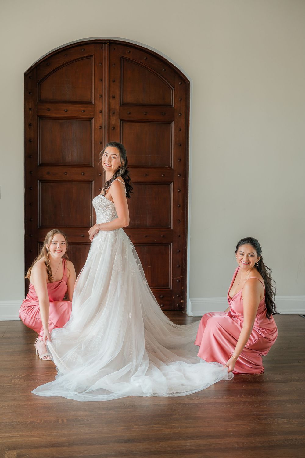 Wedding dress preparation scene with pink bridesmaid dresses against dark wooden doors.