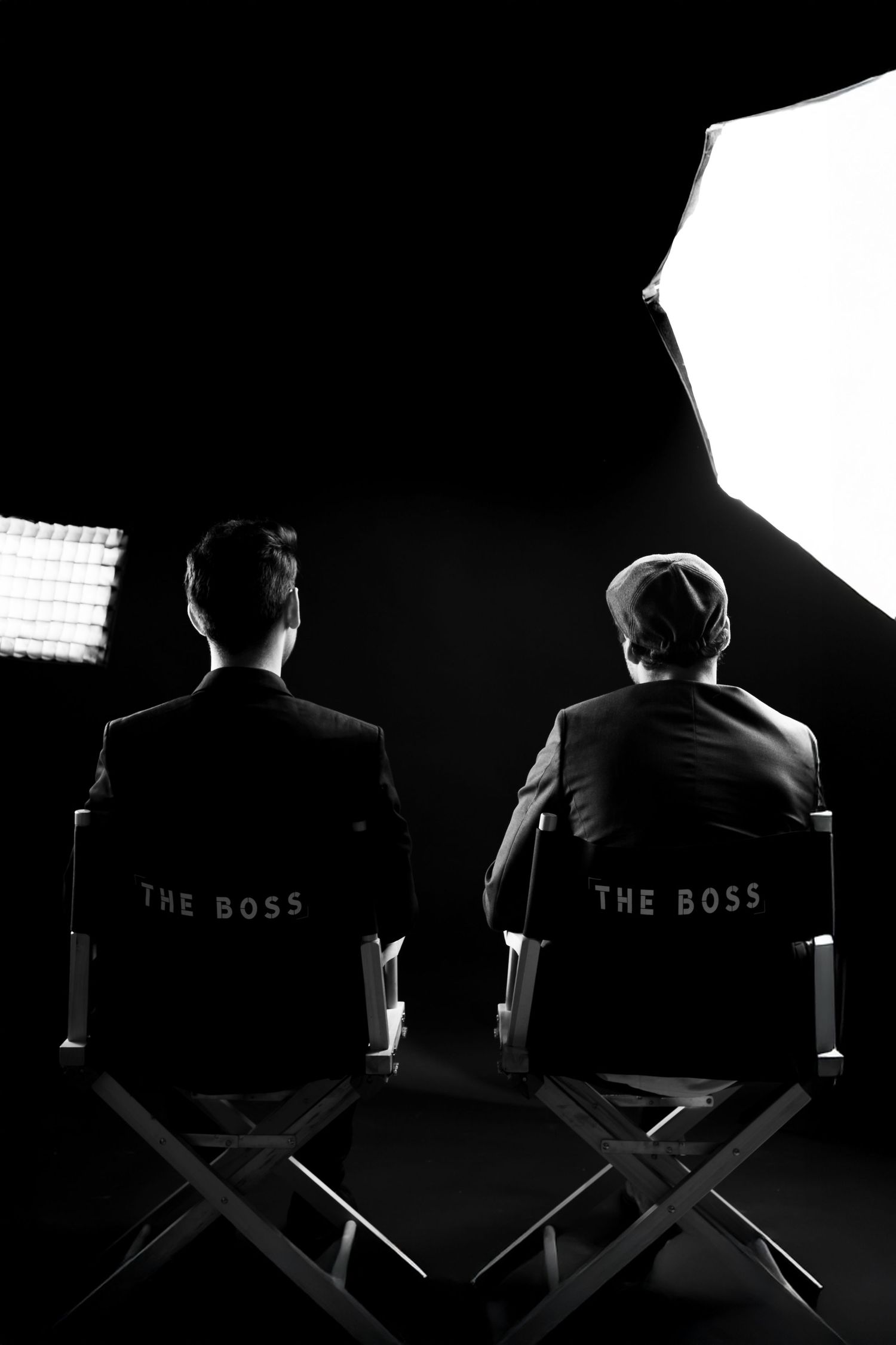 Two men in suits sit in director chairs labeled The Boss in a dramatic black and white studio setting.