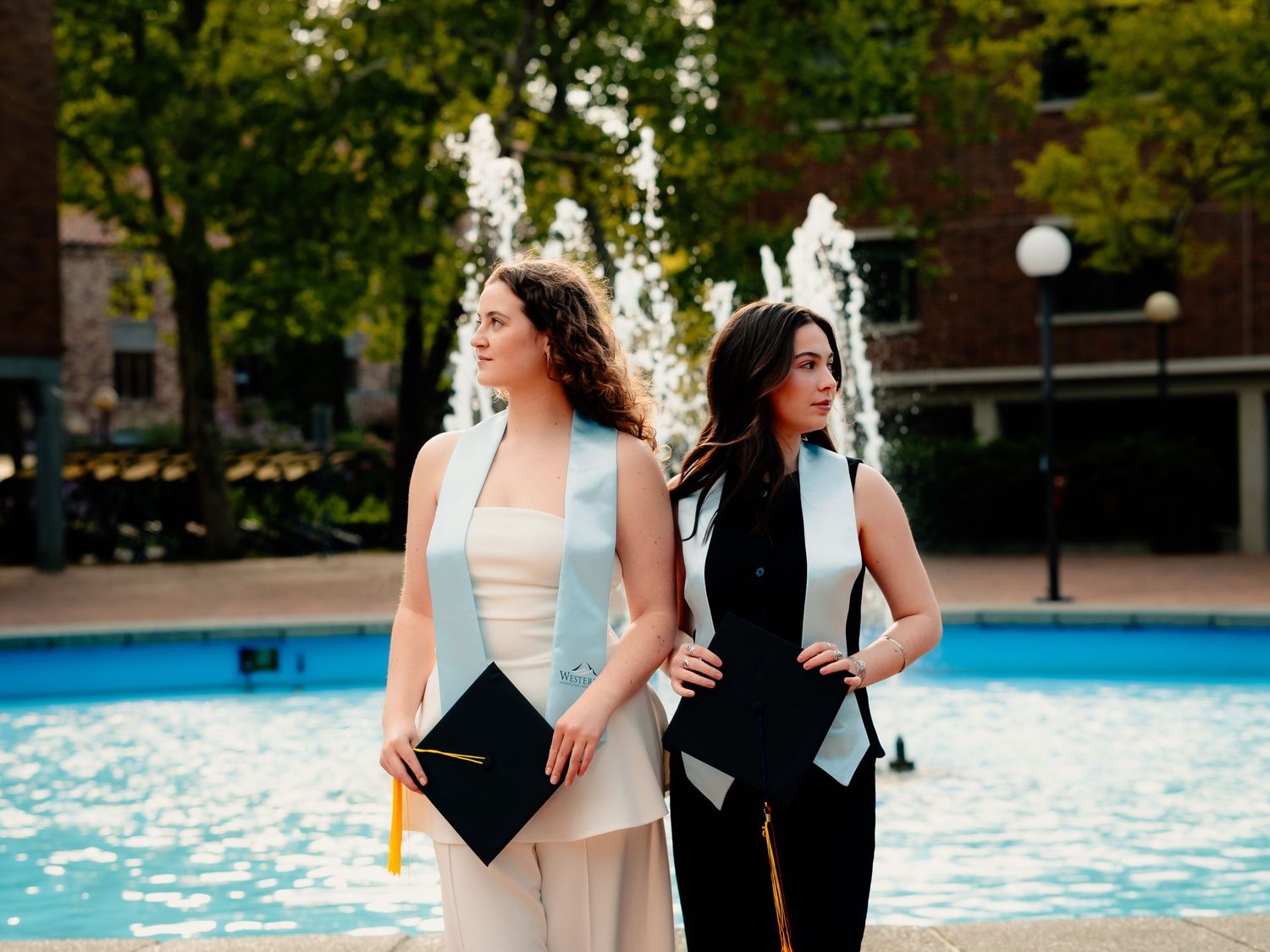 Two graduates in black caps and gowns share joyful laughter together in a sunny outdoor setting during commencement.