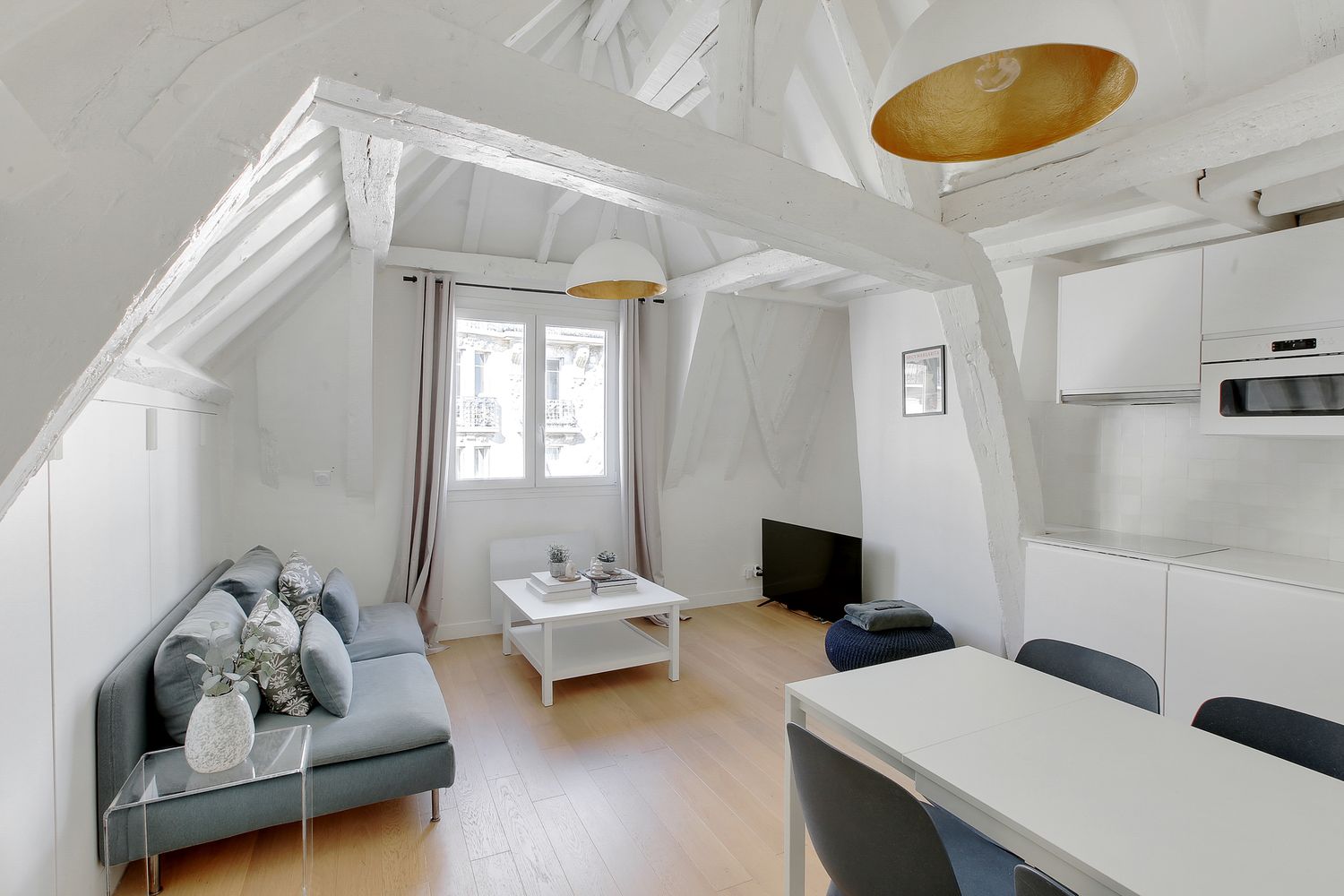 Charming attic studio apartment with white beams, gold ceiling detail, gray sofa, and compact kitchen.