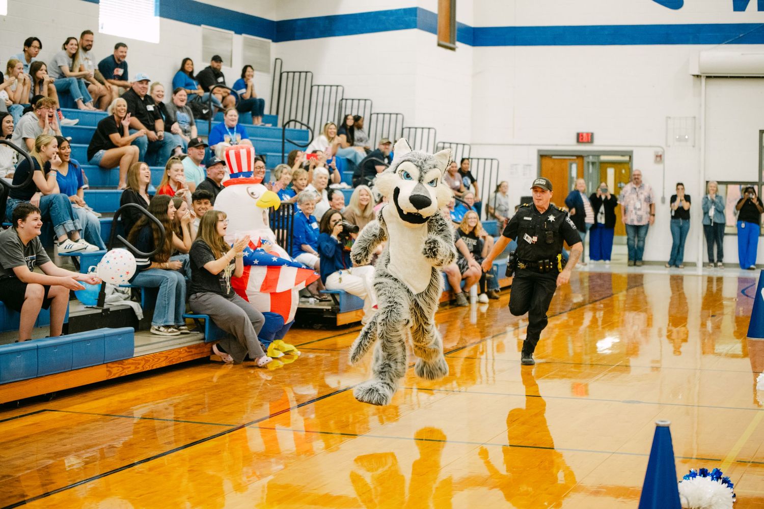 A mascot in a silver costume entertains a crowd in a school gymnasium during a sports event.