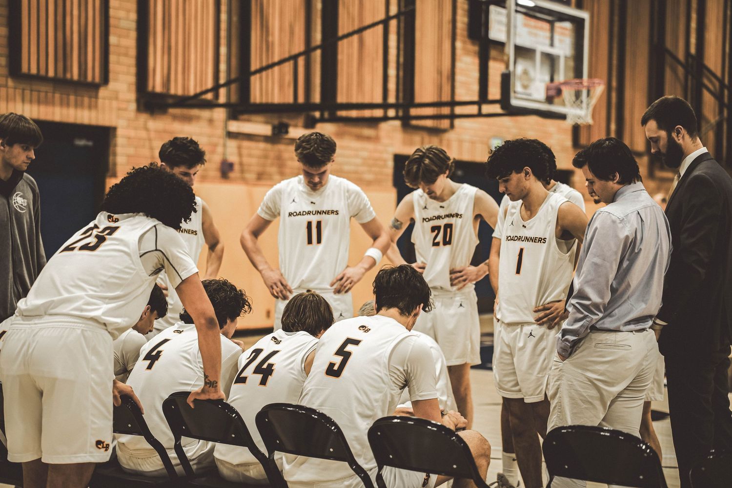 Basketball team huddles together during timeout on sidelines of indoor court.