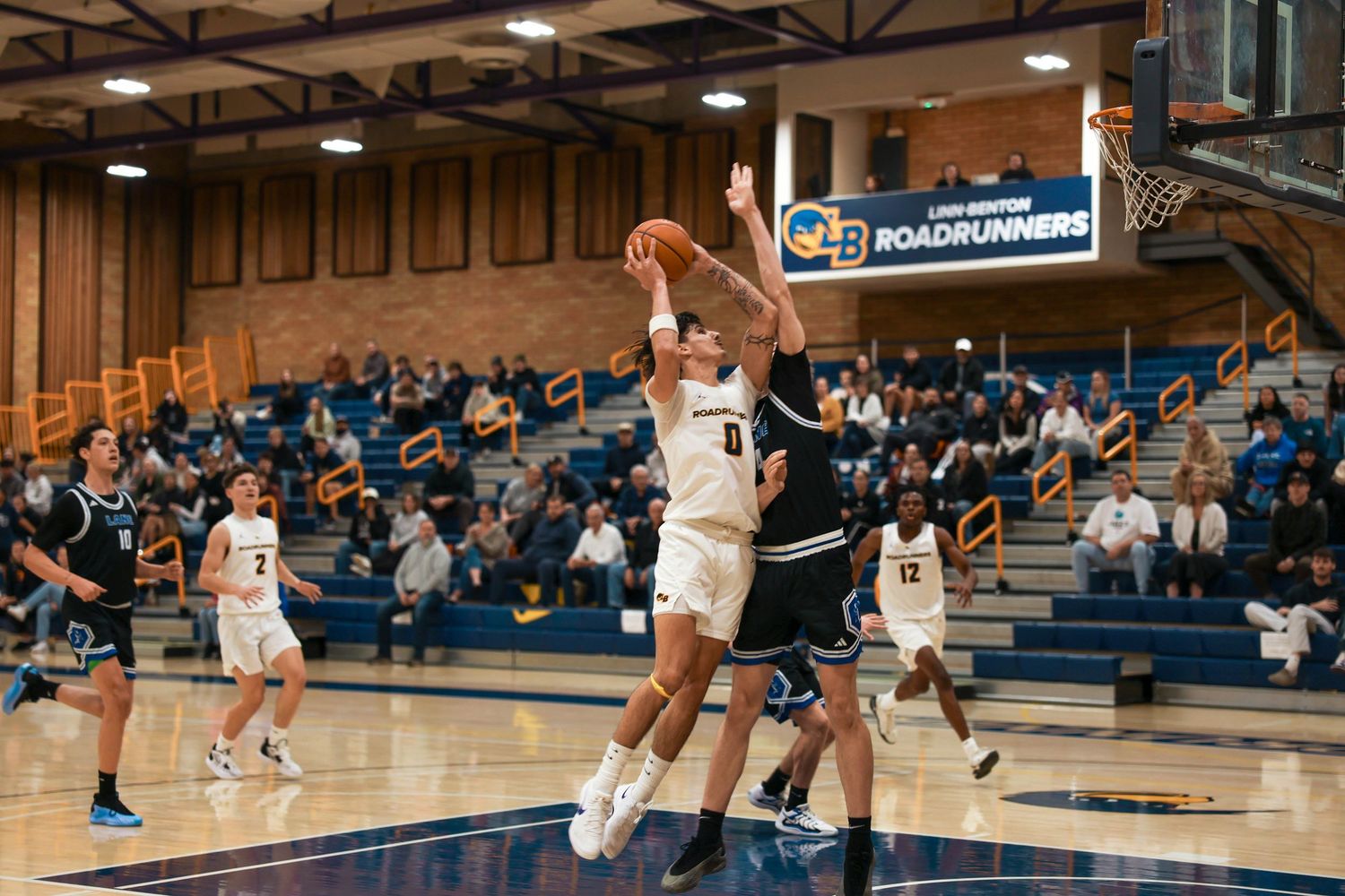 Basketball action shot showing players mid-game with spectators watching from bleachers.