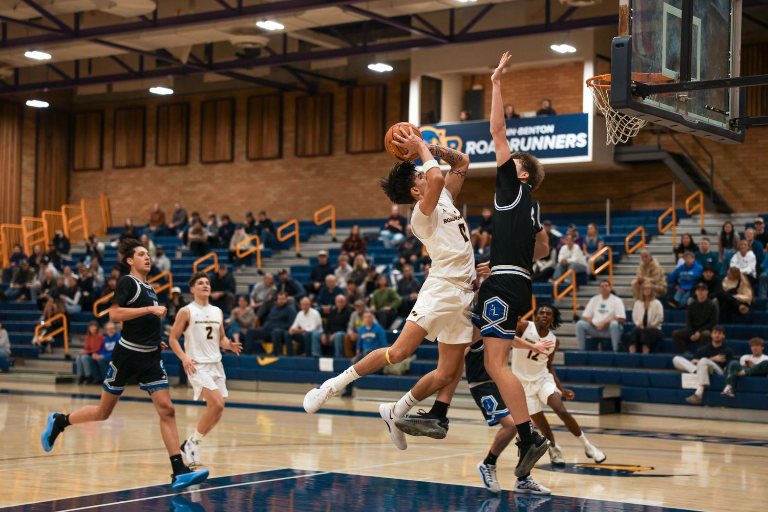 Indoor basketball game showing players in white and dark uniforms competing on court.