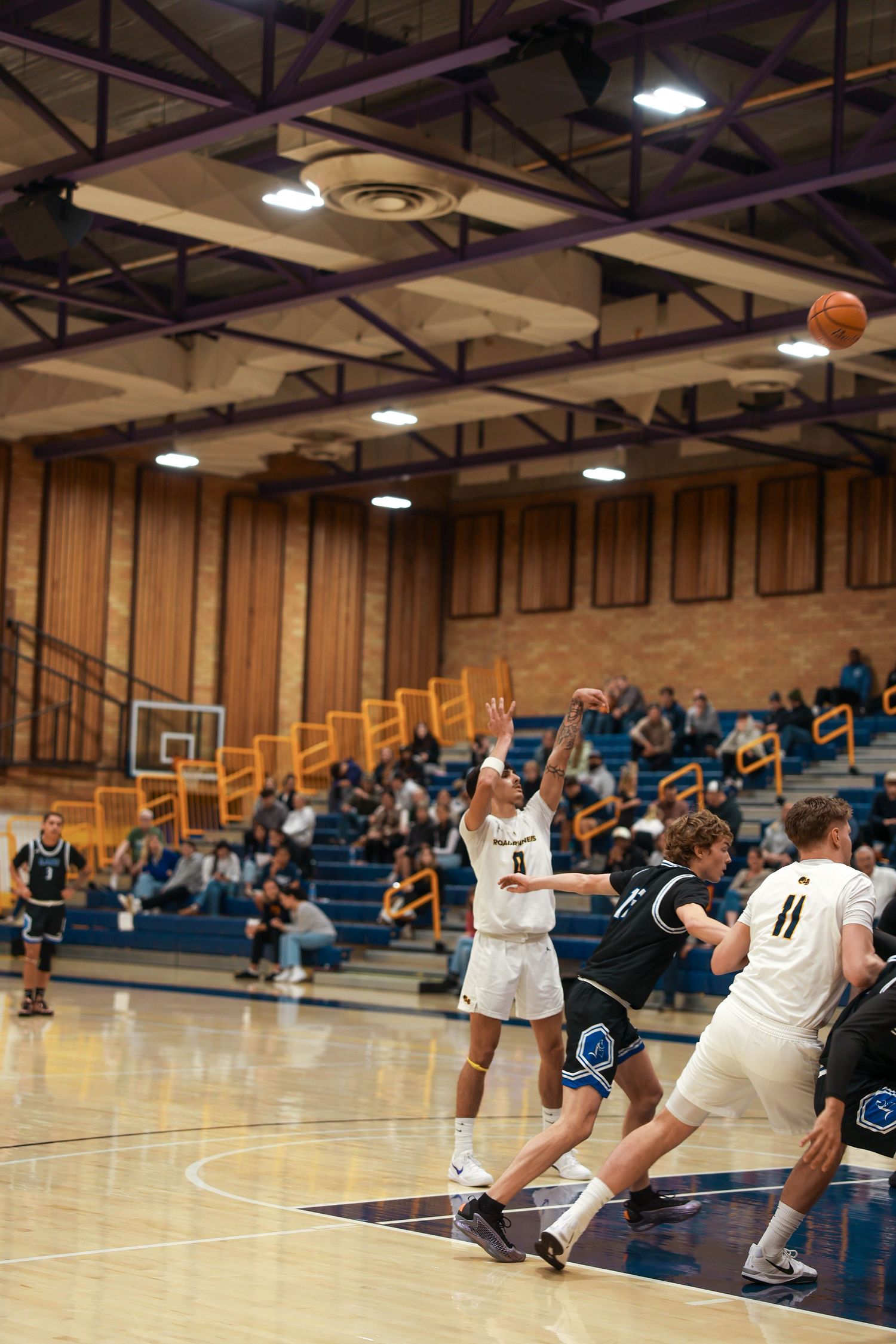 Basketball players position themselves during gameplay in gymnasium with overhead lighting.
