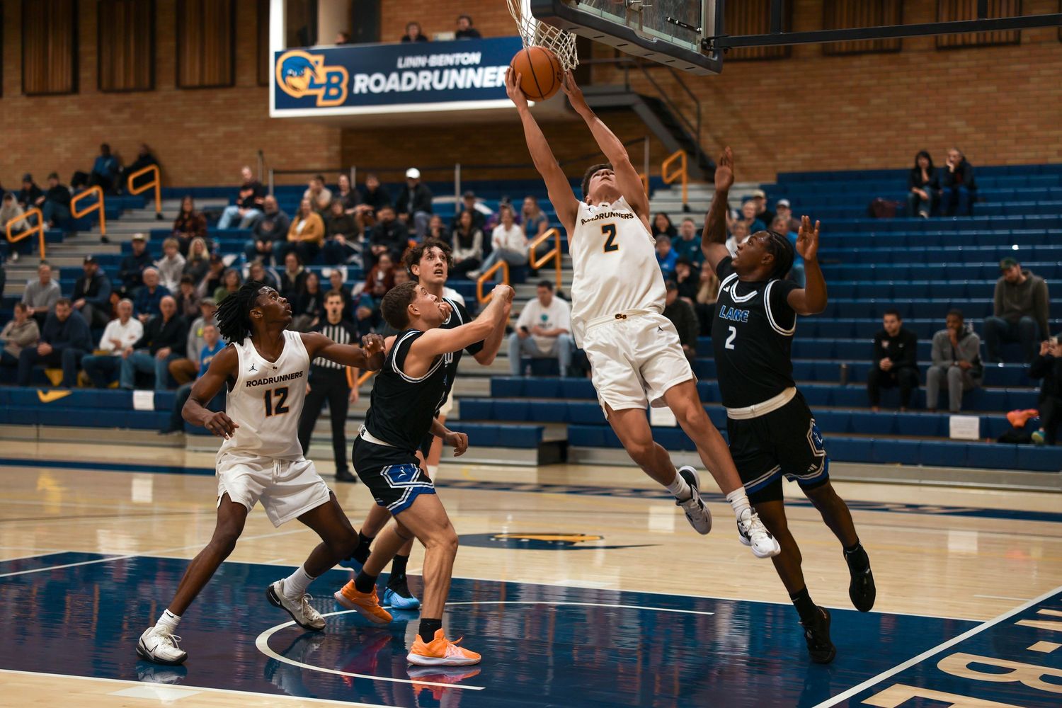 Players from opposing teams face off during a basketball match in a gymnasium with wooden walls.