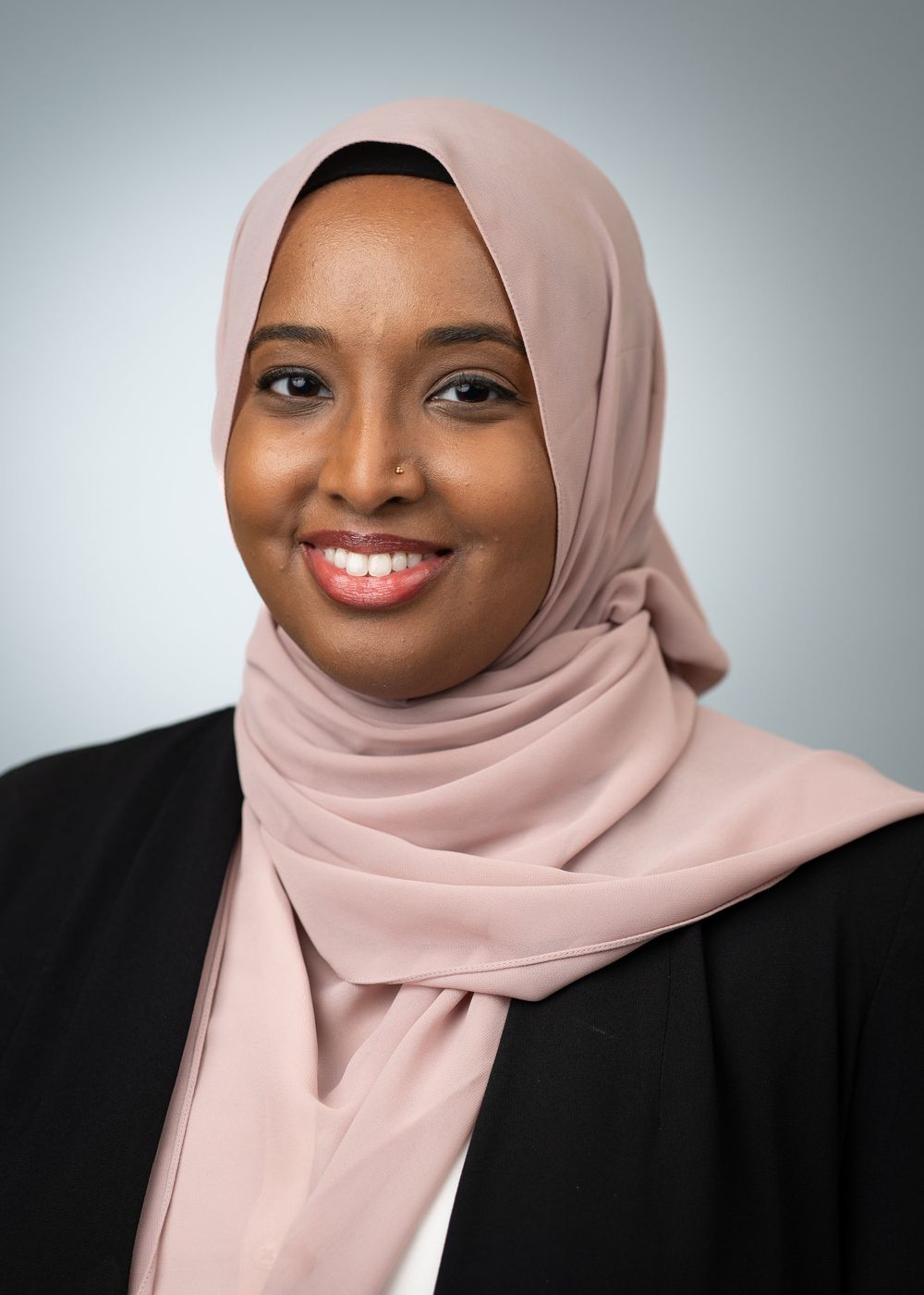 A person wearing a pink hijab and black blazer smiles warmly against a light gray background.