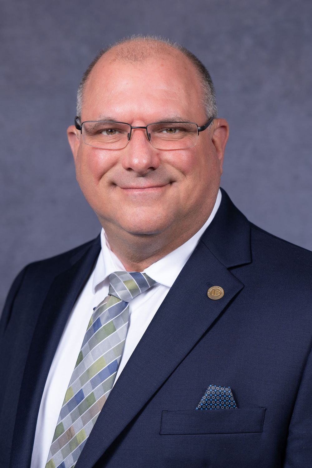 Professional headshot of someone in a dark business suit with striped tie and glasses against a blue backdrop.