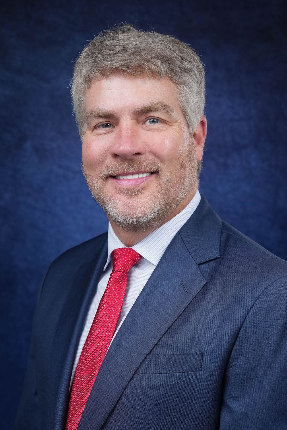 Professional headshot of someone wearing a dark suit and red tie against a blue background.