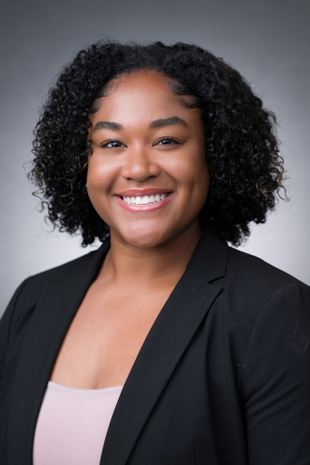 Professional headshot portrait series showing bright smile and natural curly hair against gray background wearing black blazer.