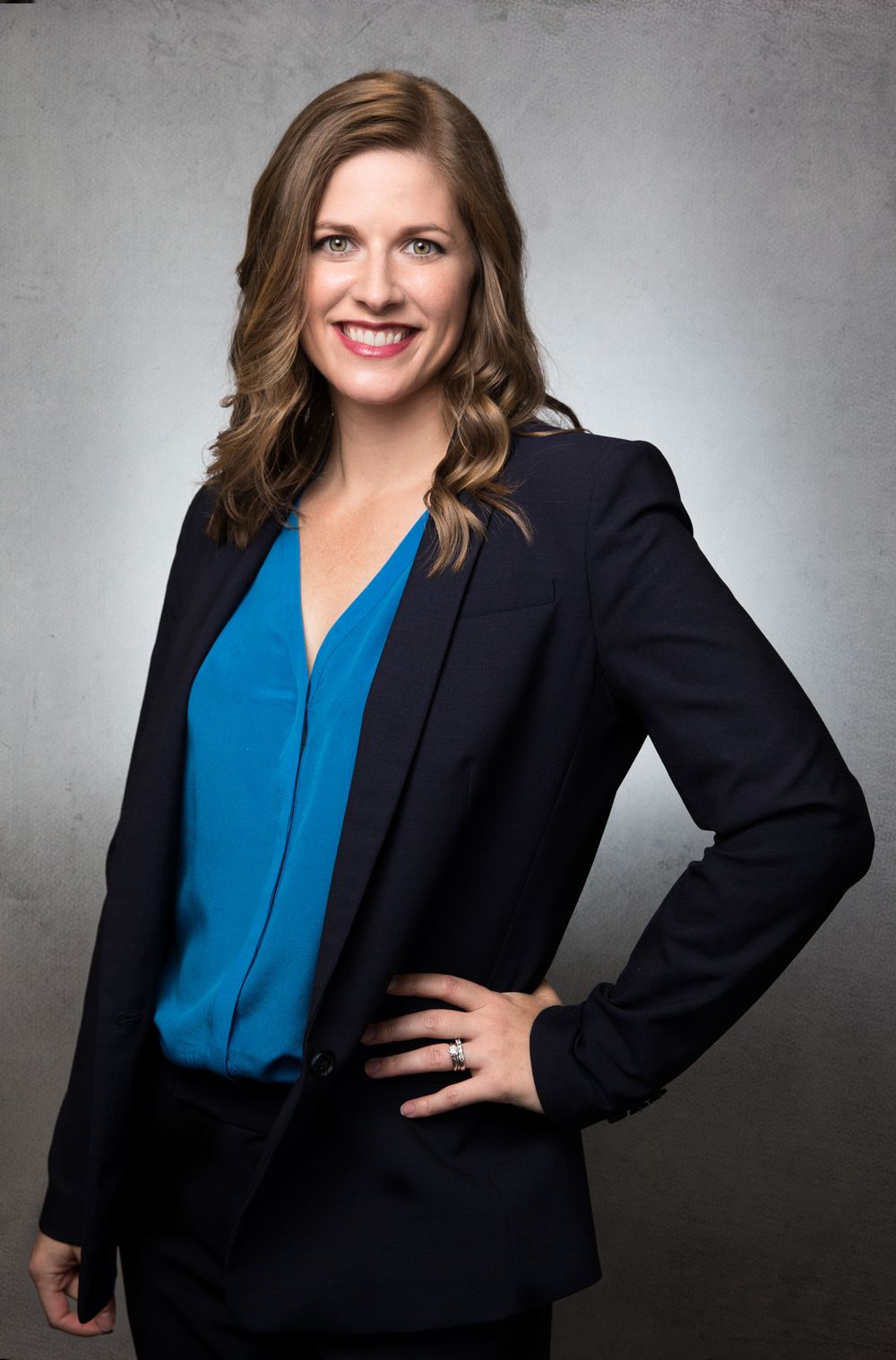 Professional portrait of someone in a navy blazer and blue blouse against a gray studio backdrop.