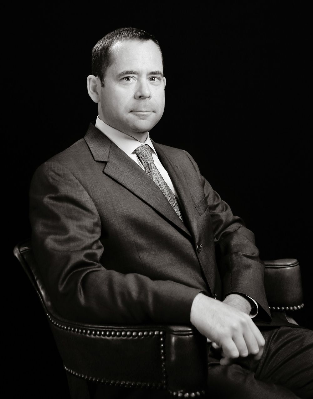 Professional black and white portrait photograph showing a person in a dark suit seated in a studded leather chair.