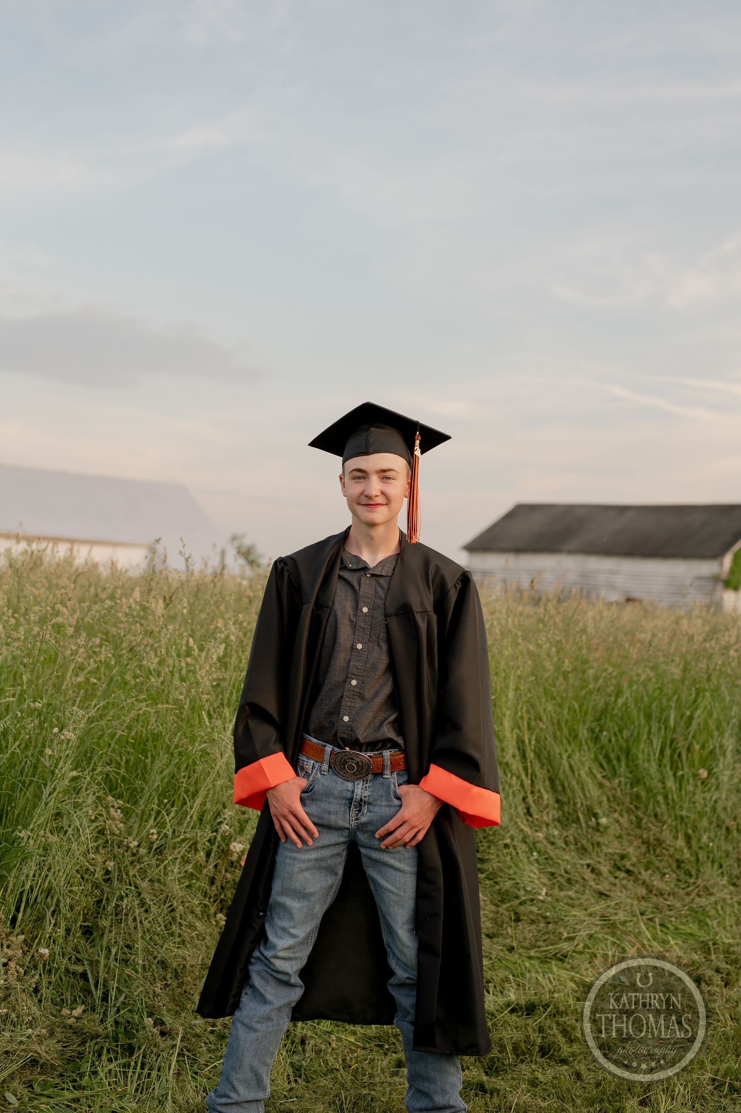A graduate in black cap and gown poses in a rural field with barn in background.