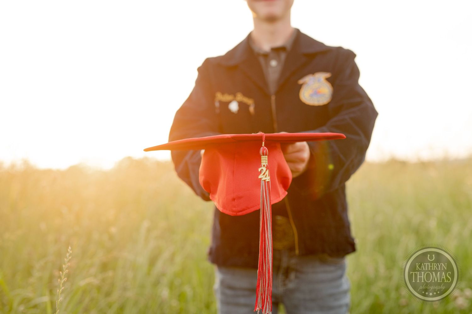 Red graduation cap and tassel backlit by golden sunlight in field setting.