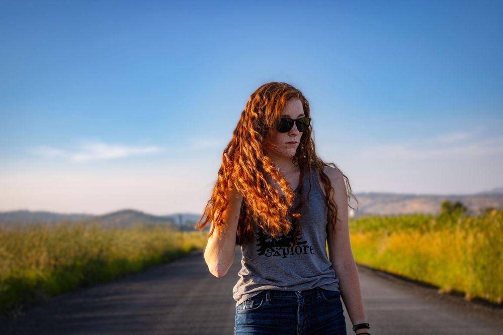 redhaired girl stands facing slightly right with sunglasses on and hand in hair.  she is on a rural road with clear sky and hills behind her