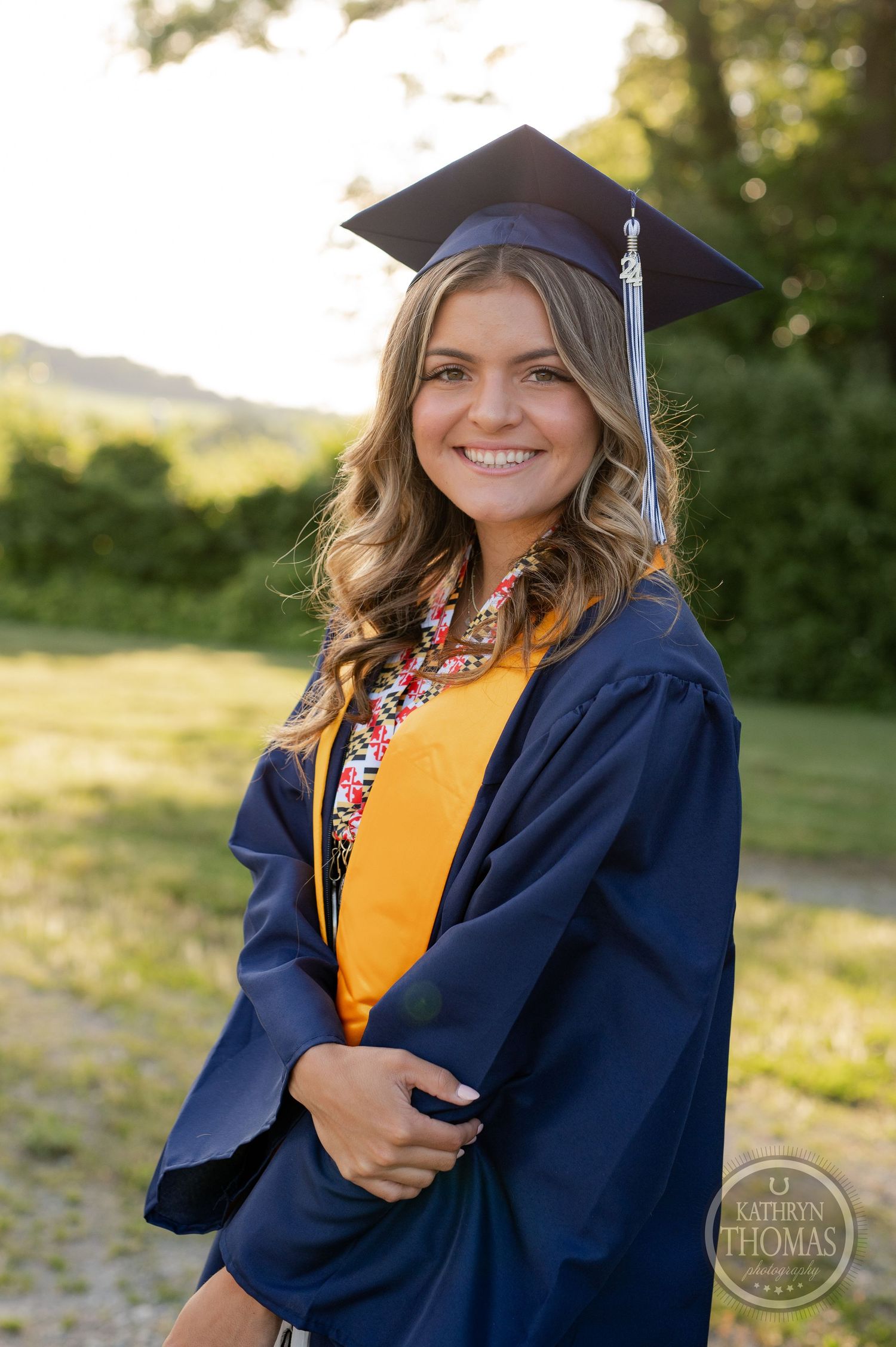 Graduate in blue cap and gown with yellow stole poses outdoors during golden hour.