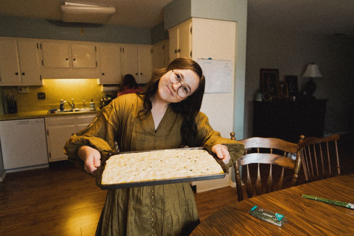 Person in yellow sweater and green dress holding a baking sheet in a kitchen.
