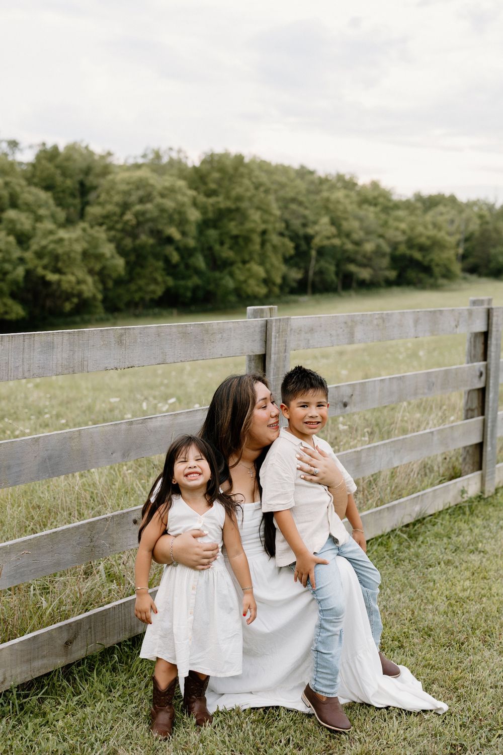 Family shares joyful moment together by rustic wooden fence in grassy field on cloudy day.