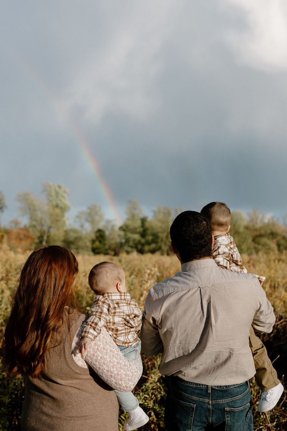 A family admires a rainbow together in a field during golden hour with dramatic cloudy skies.