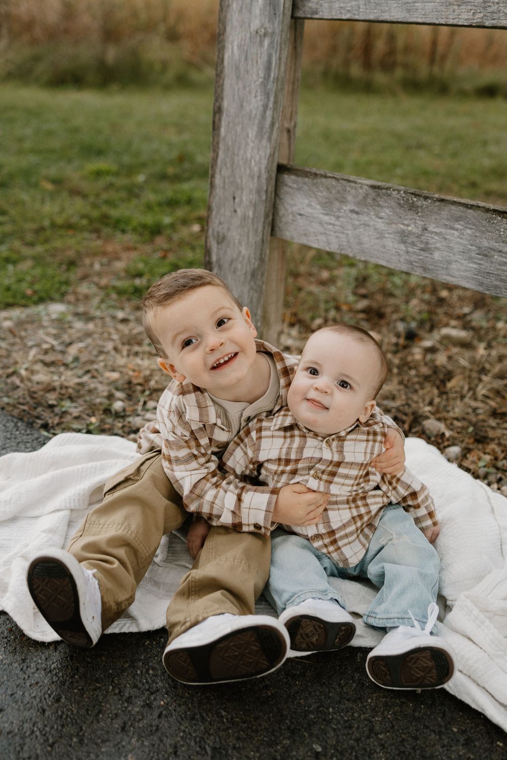 Two young children in matching plaid shirts sit together on a white blanket against a wooden fence outdoors.