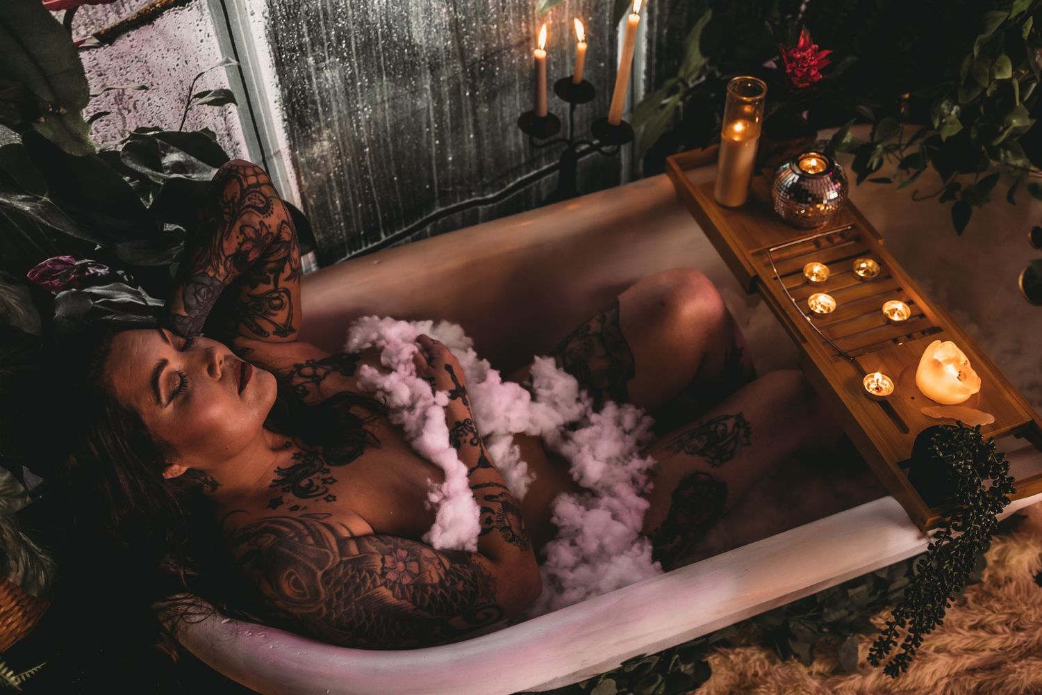 Plus size tattooed woman laying in clawfoot tub with candles lit and bubbles during empowering boudoir photoshoot