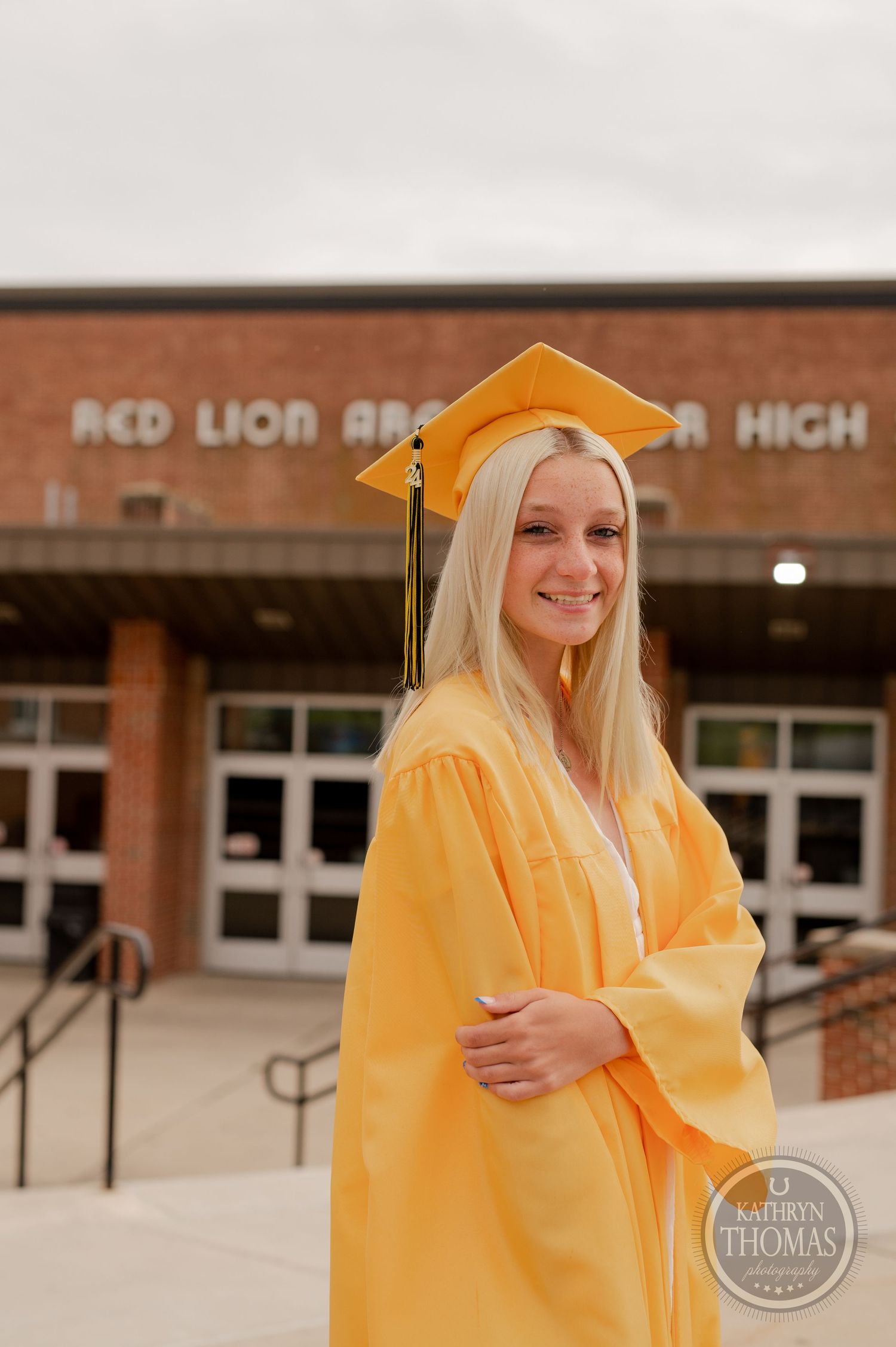 Graduate in bright yellow cap and gown smiles brightly outside school entrance.