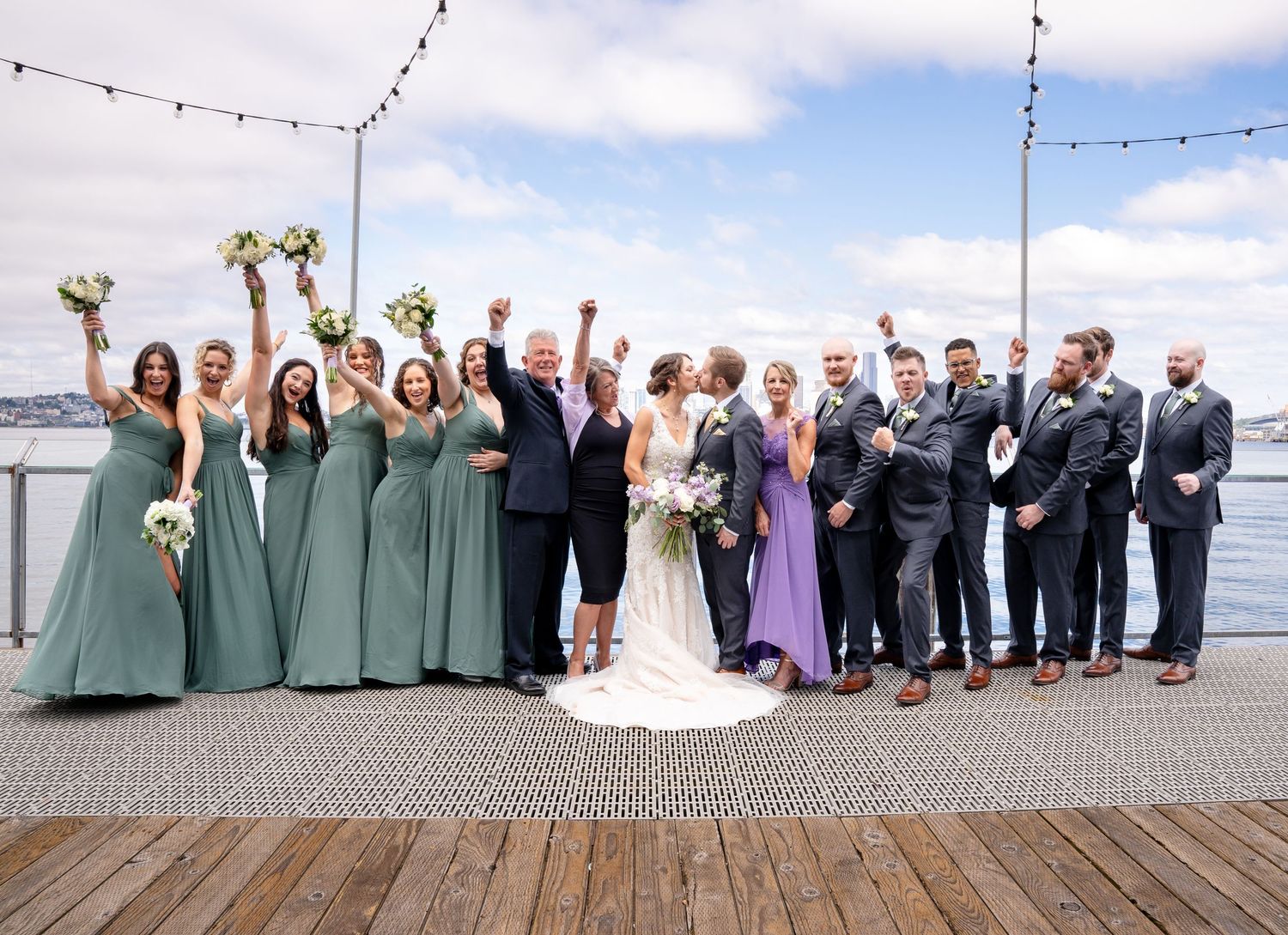 Wedding party in sage green and gray attire celebrates on a waterfront dock under string lights and blue sky.