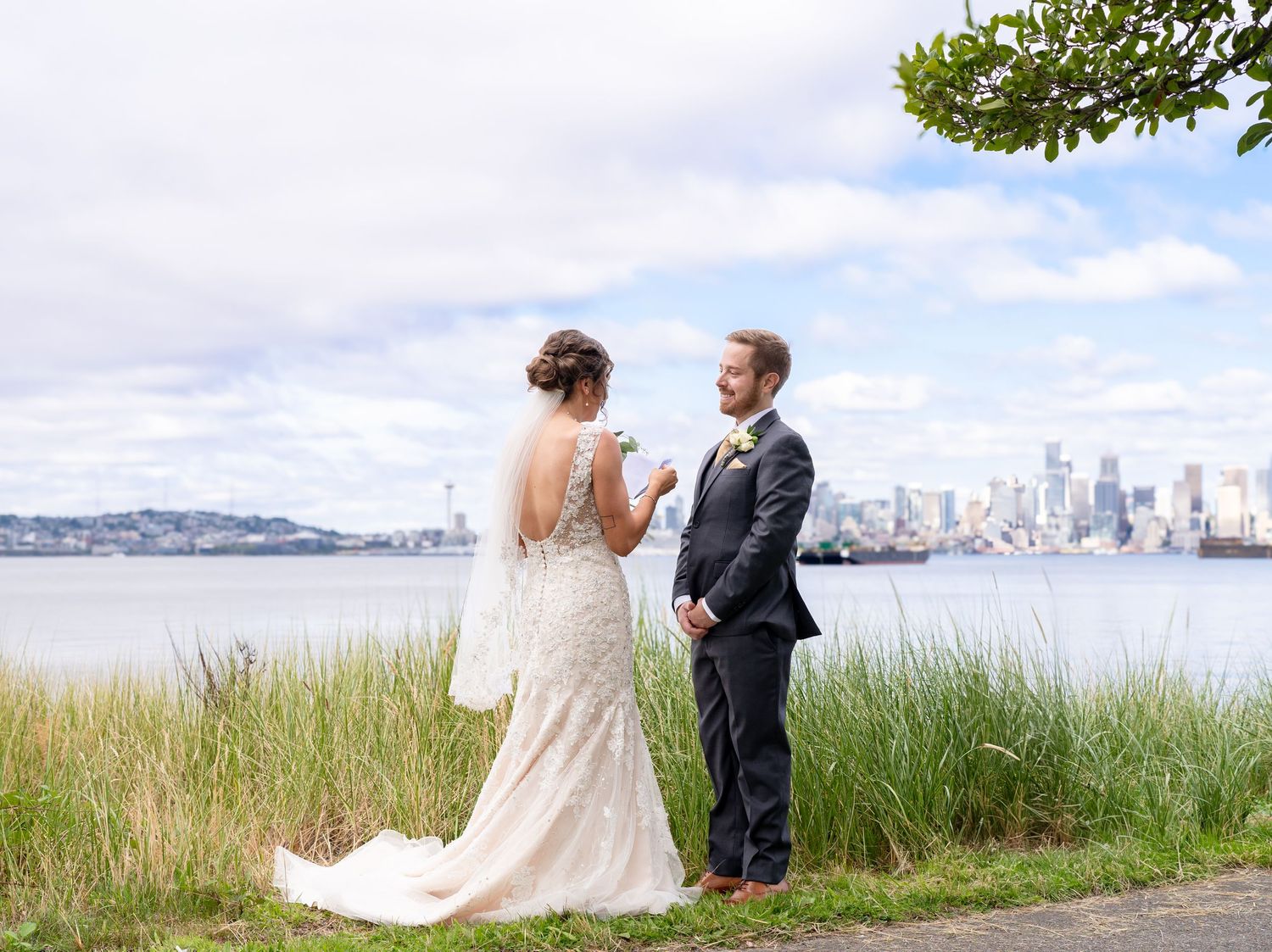 A bride and groom share a moment on a grassy hilltop overlooking a city skyline across the water.