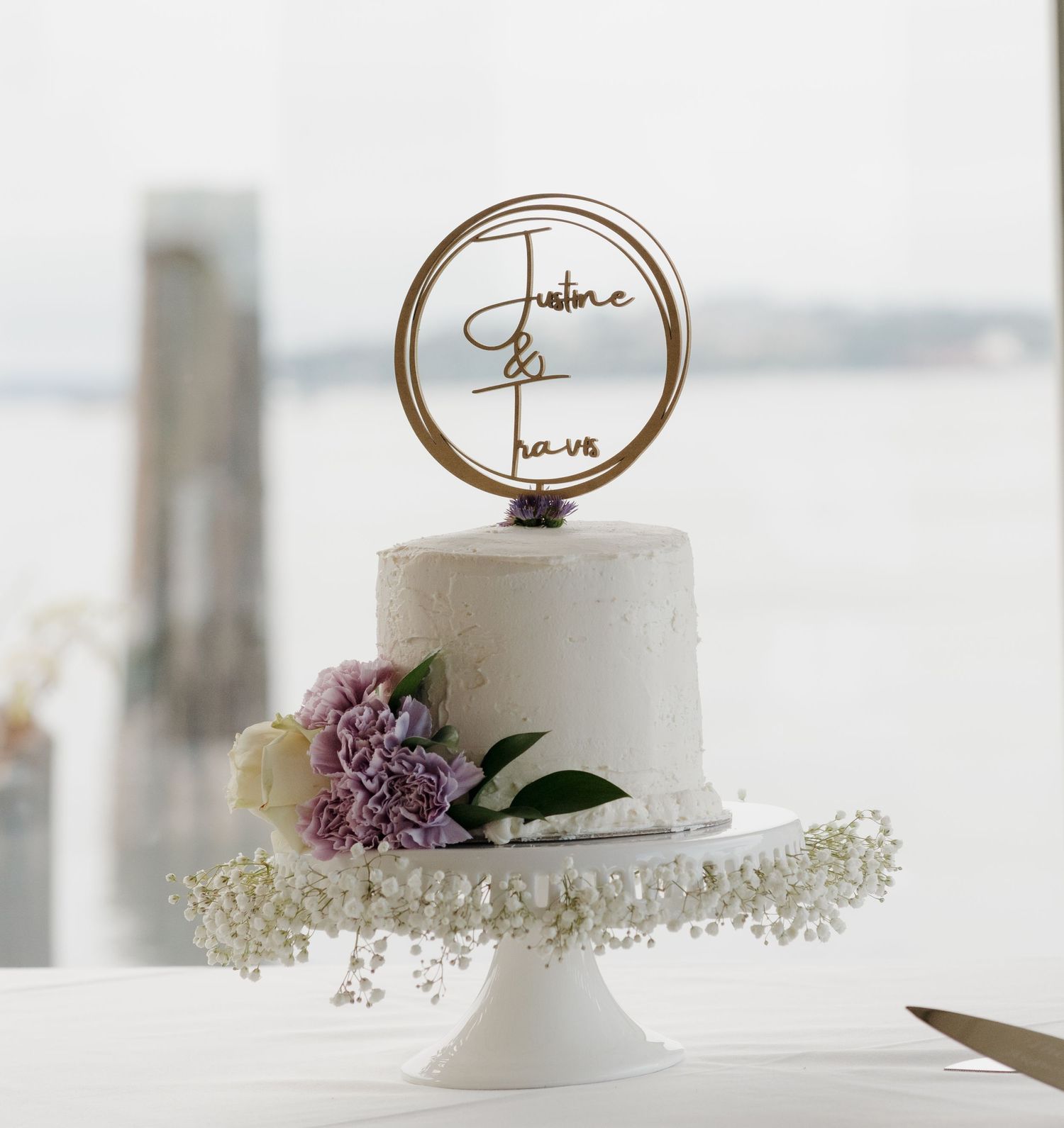 Elegant white wedding cake with a round 'forever' topper and purple flower accent on white pedestal stand.