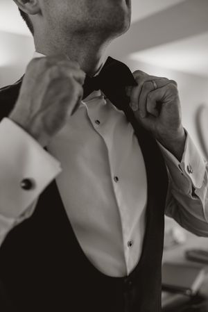 Close-up black and white photo of someone adjusting a black bow tie while wearing a formal tuxedo and white dress shirt.
