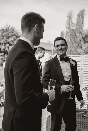 A group of men in formal black suits and ties chat and drink champagne at an outdoor event.