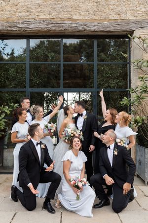 Wedding party in formal attire shares laughs and drinks while sitting against stone wall at outdoor venue.